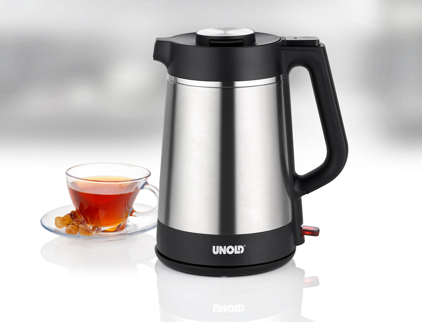 Der Unold Blitzkocher Thermo 1,5 L Wasserkocher aus Edelstahl mit schwarzem Griff steht auf einer reflektierenden Oberfläche neben einer Glastasse Tee und einem Teller Kandiszucker.