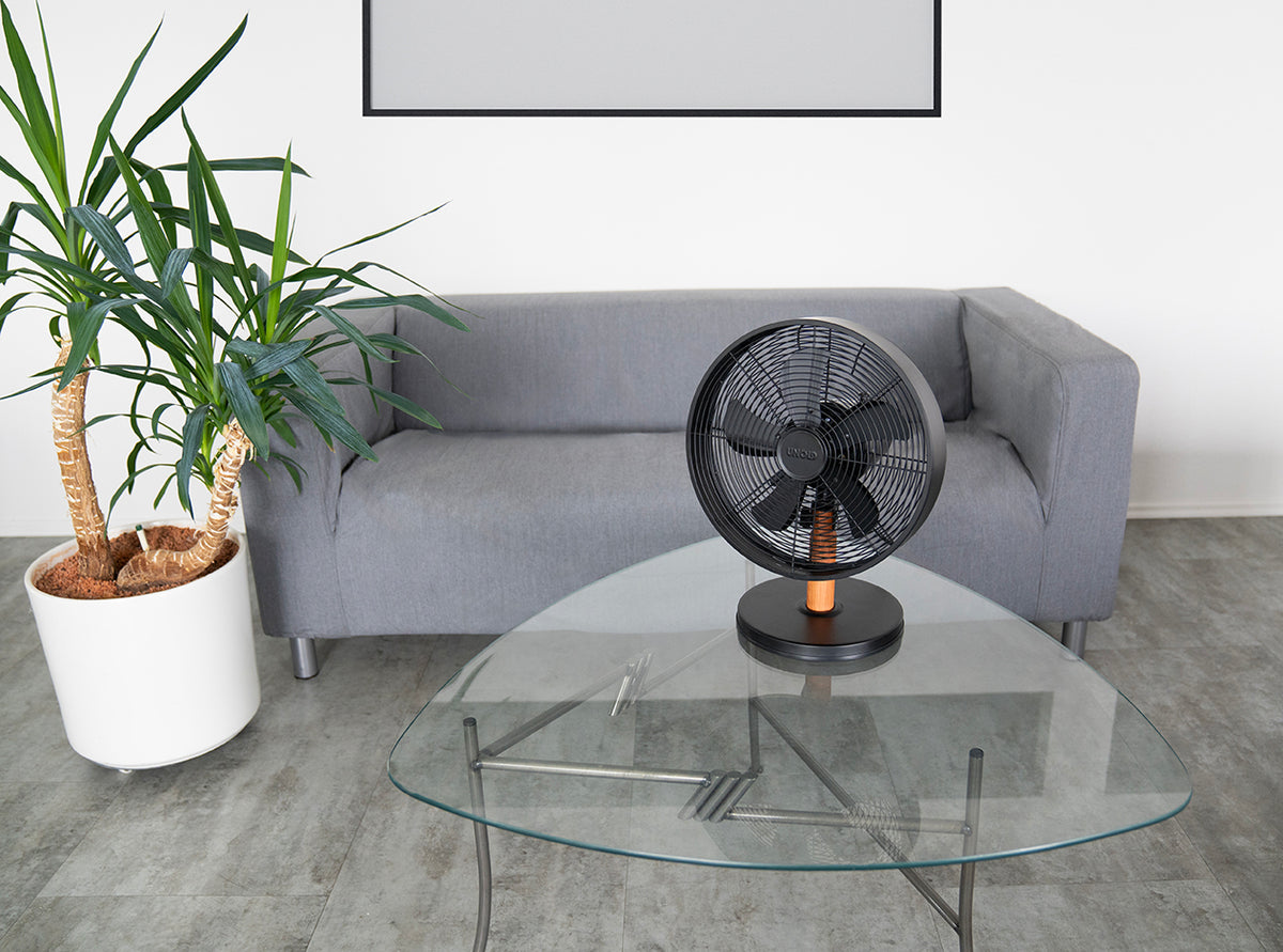 Der Unold Tischventilator Black Steel steht auf einem Couchtisch aus Glas vor einem grauen Sofa, links davon steht eine große Topfpflanze, alles in einem modernen, minimalistischen Raum mit grauem Boden.