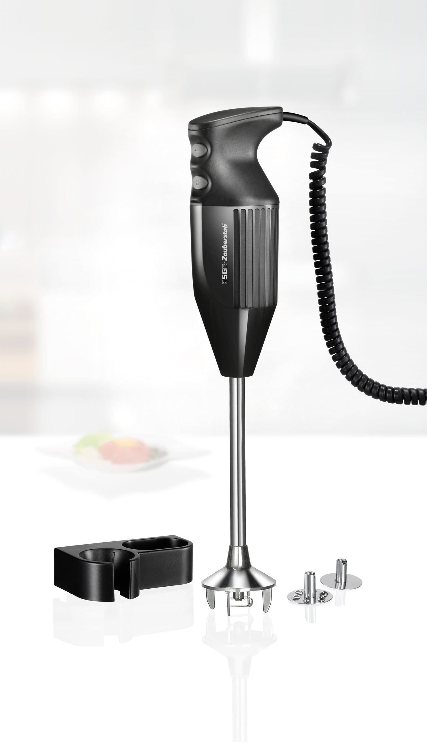 Ein schwarzer Stabmixer mit Spiralkabel steht aufrecht auf einer weißen Fläche, begleitet von einer Wandhalterung und drei metallischen Mixaufsätzen. Im Hintergrund ist eine unscharfe Küchenszene zu sehen.