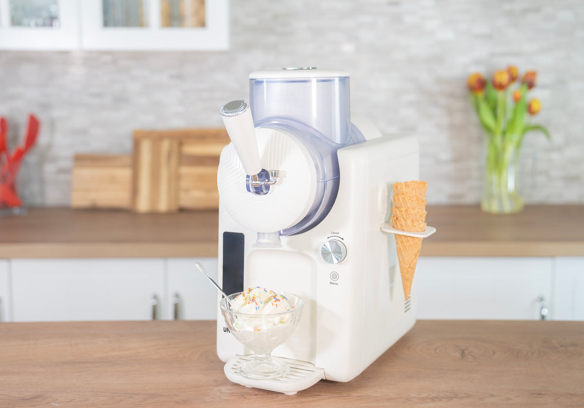 Eine Eismaschine aus Edelstahl, umgeben von Schalen mit buntem Eis und Sorbet, garniert mit Früchten, Nüssen und Beeren, vor einem hellen, unscharfen Küchenhintergrund.