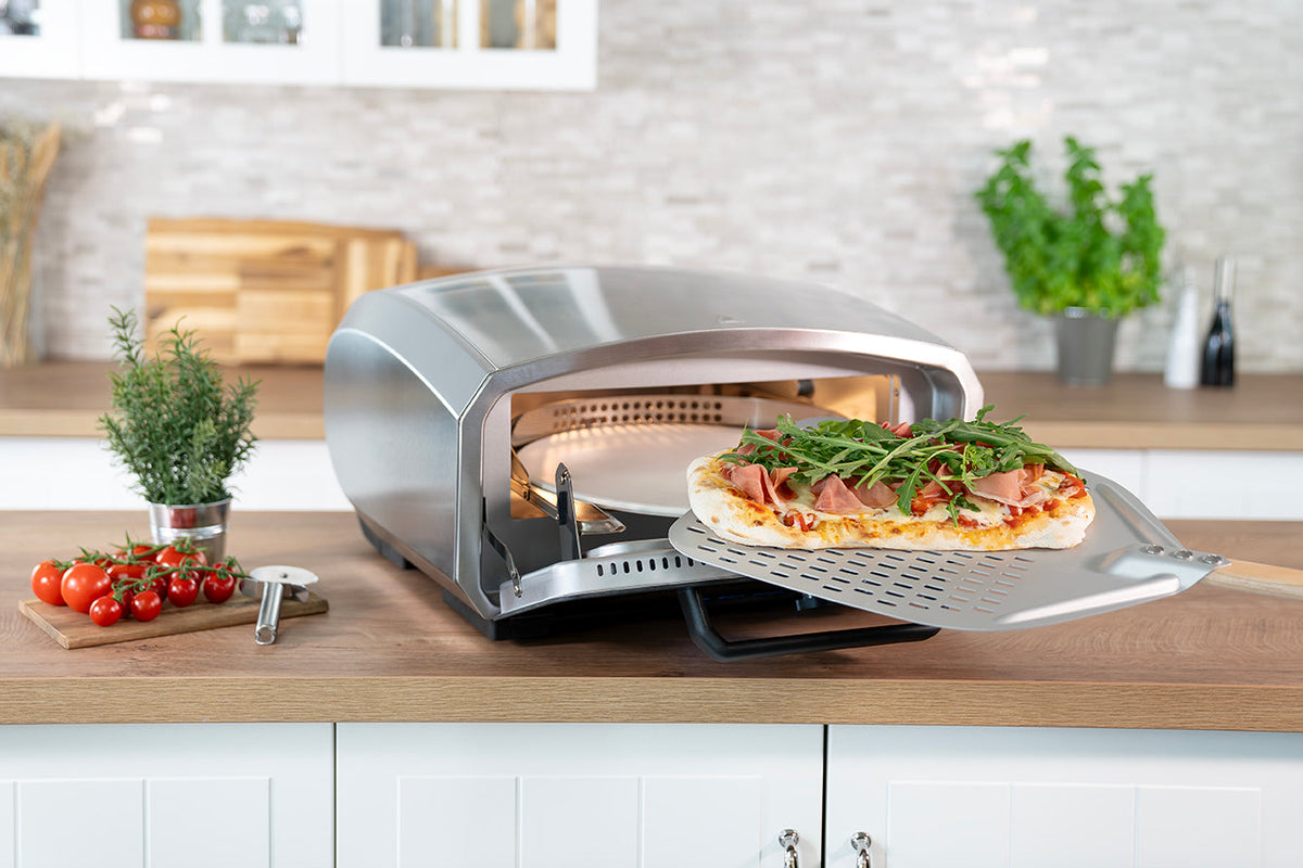 Ein Pizzaofen steht auf einem Küchentisch, daneben stehen frische Kräuter, Tomaten, ein Pizzaschneider und eine fertige Pizza auf einer Schiene. Im Hintergrund sind weiße Schränke und Pflanzen zu sehen.
