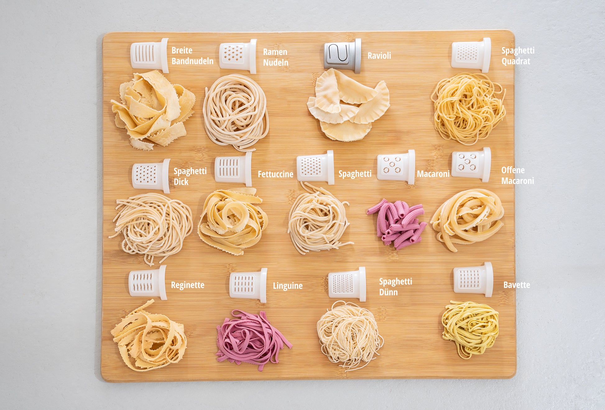 Die Unold Nudelmaschine Nora verfügt über 15 Nudelsorten, jede mit deutscher Beschriftung und den entsprechenden Aufsätzen auf einem Holzbrett angeordnet, und zeigt Formen wie Spaghetti, Fettuccine, Ravioli und Makkaroni.