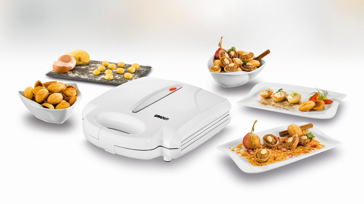 Der Unold Waffelautomat Zaubernuss steht auf einer weißen Fläche, umgeben von Tellern mit Gebäck, gebackenen Desserts, rohen Teigbällchen auf schwarzem Schiefer sowie Eiern und Mehl.