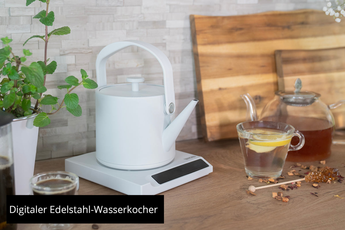 Der Unold Blitzkocher Clara, ein weißer Wasserkocher auf digitalem Sockel, steht auf einer Holztheke mit einer Glasteekanne, Zitronentee, Minze, Teeblättern und zwei Schneidebrettern. Text: Digitaler Edelstahl-Wasserkocher.