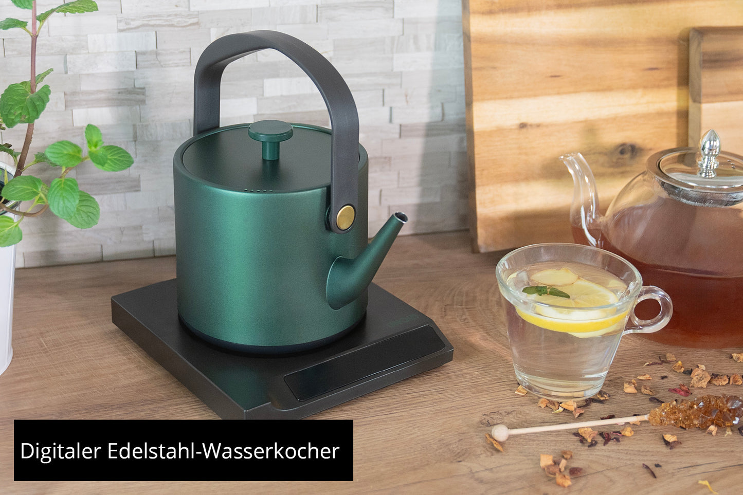 Der Unold Blitzkocher Jade, ein grüner digitaler Edelstahl-Wasserkocher, steht auf einem schwarzen Sockel neben einer Glasteekanne, einer Tasse Zitronentee, losen Teeblättern und einer Topfpflanze. Der Text lautet: „Digitaler Edelstahl-Wasserkocher“.