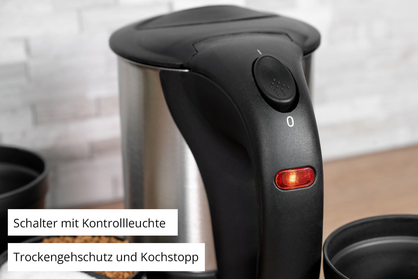 Eine Nahaufnahme des schwarzen Griffs des Unold Reiseblitzkochers Edelstahl 0,5 l zeigt eine rote Kontrollleuchte. Auf den deutschen Etiketten steht: „Schalter mit Kontrollleuchte“ sowie „Trockengehschutz und Kochstopp“. Daneben liegen Teile von Tassen.