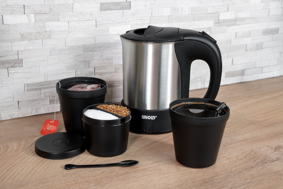 Der Unold Reiseblitzkocher Edelstahl 0,5 l steht auf einer Holzunterlage neben drei schwarzen Behältern mit Teebeuteln, Zucker und Kaffeepulver sowie einem schwarzen Löffel und einer Tasse Kaffee.