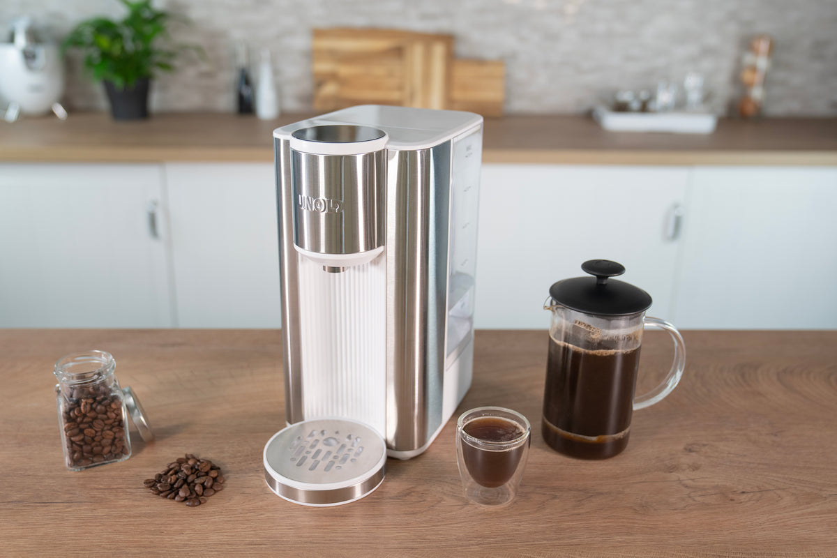 Der Unold Express Heißwasserbereiter Ava in Silber und Weiß steht auf einer Holzarbeitsplatte neben einer mit Kaffee gefüllten French Press aus Glas, einer kleinen Kaffeetasse, einem Glas Bohnen und losen Bohnen, alles in einer modernen Küche.