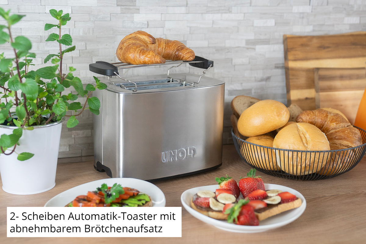 Der Unold Toaster Thommy mit herausnehmbarem Rost toastet ein Croissant auf der Küchentheke neben einer Topfpflanze, Brötchen, einem Schneidebrett und Tellern mit Toast, Obst und Aufstrichen.