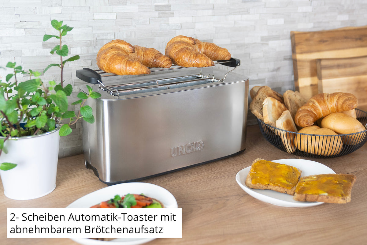 Der Unold Toaster Tom aus Edelstahl steht mit zwei Croissants auf der Küchentheke. In der Nähe stehen eine Topfpflanze, ein Brotkorb, ein Teller mit Toast und Ei, ein Schneidebrett mit Croissants und ein deutscher Text.
