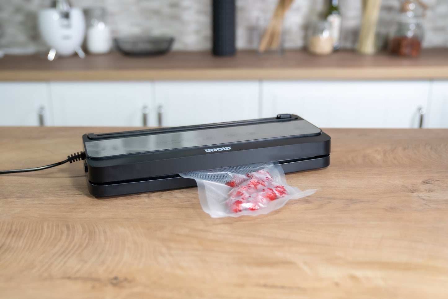 Der Unold Vakuumierer Food Saver Black verschließt einen mit roten und weißen Bonbons gefüllten Plastikbeutel auf einer hölzernen Küchentheke, im Hintergrund sind unscharfe Küchenutensilien und Gläser zu sehen.