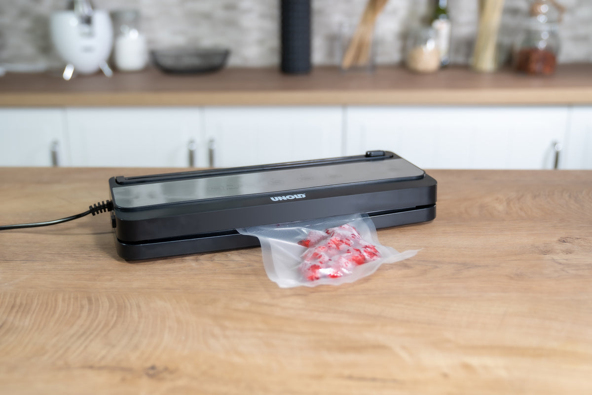 Der Unold Vakuumierer Food Saver Black verschließt einen mit roten und weißen Bonbons gefüllten Plastikbeutel auf einer hölzernen Küchentheke, im Hintergrund sind unscharfe Küchenutensilien und Gläser zu sehen.