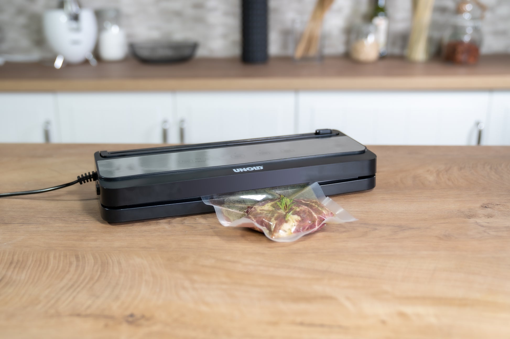 Der Unold Vakuumierer Food Saver Black steht auf einer Küchentheke und verschließt vor einem modernen Küchenhintergrund einen mit rohem Fleisch und Kräutern gefüllten Beutel.