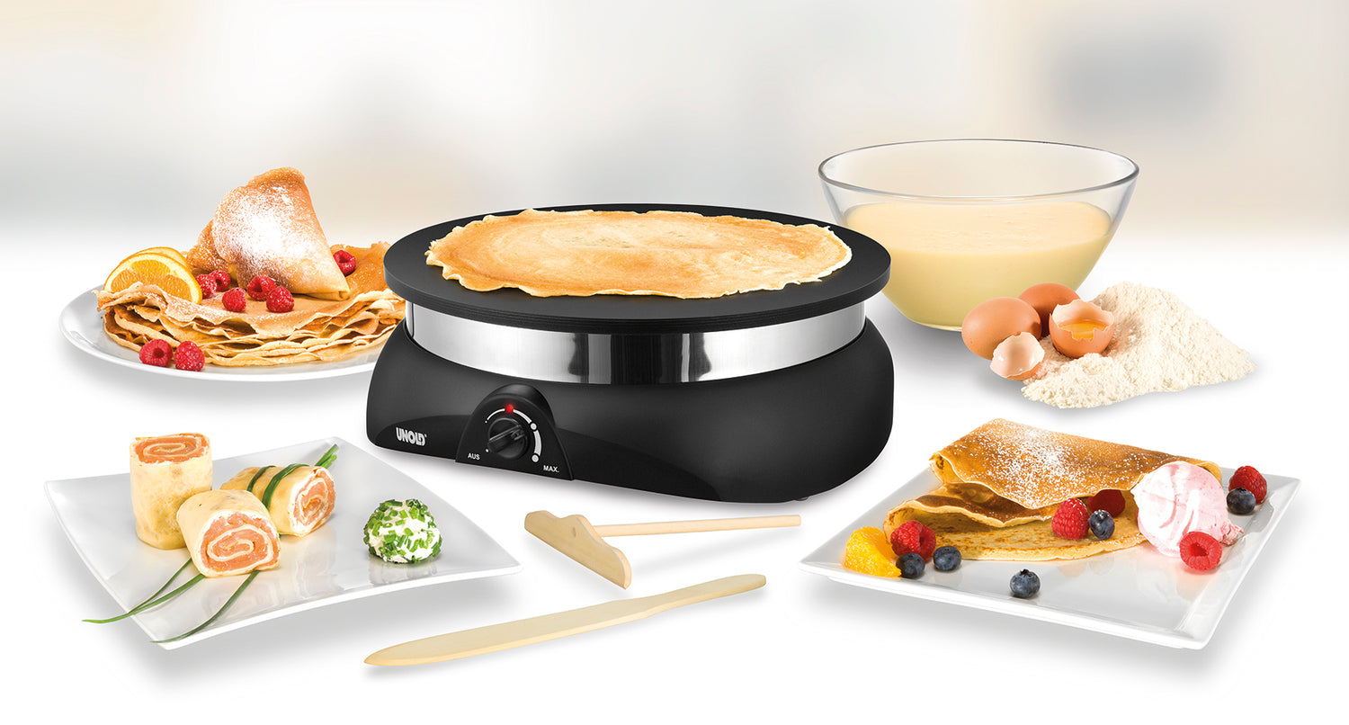 Ein elektrischer Crêpe-Maker, umgeben von Tellern mit Crêpes mit verschiedenen Füllungen, einer Schüssel mit Teig, Eiern und Mehl. Davor stehen Holzutensilien, der Hintergrund ist leicht verschwommen.