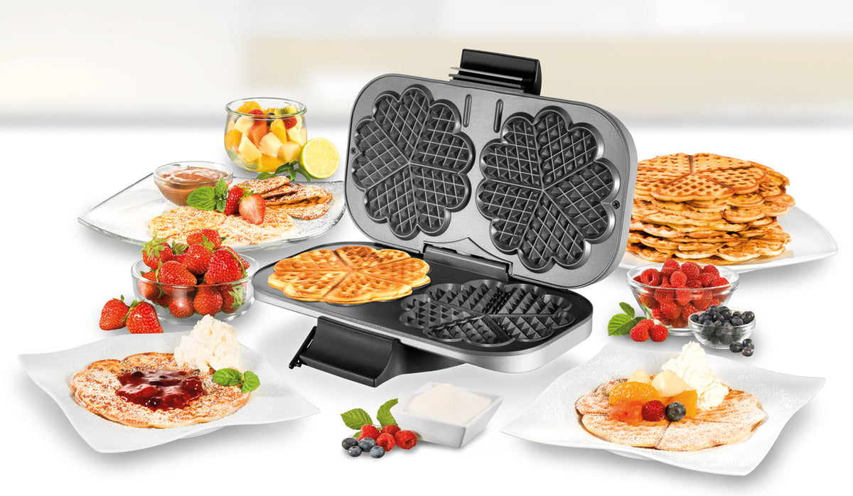 Der Doppel-Waffelautomat von Unold steht geöffnet auf einem weißen Tisch, umgeben von Tellern mit herzförmigen Waffeln, garniert mit Früchten, Sirup und Schlagsahne, sowie Schüsseln mit frischen Erdbeeren, Blaubeeren und Obstsalat.