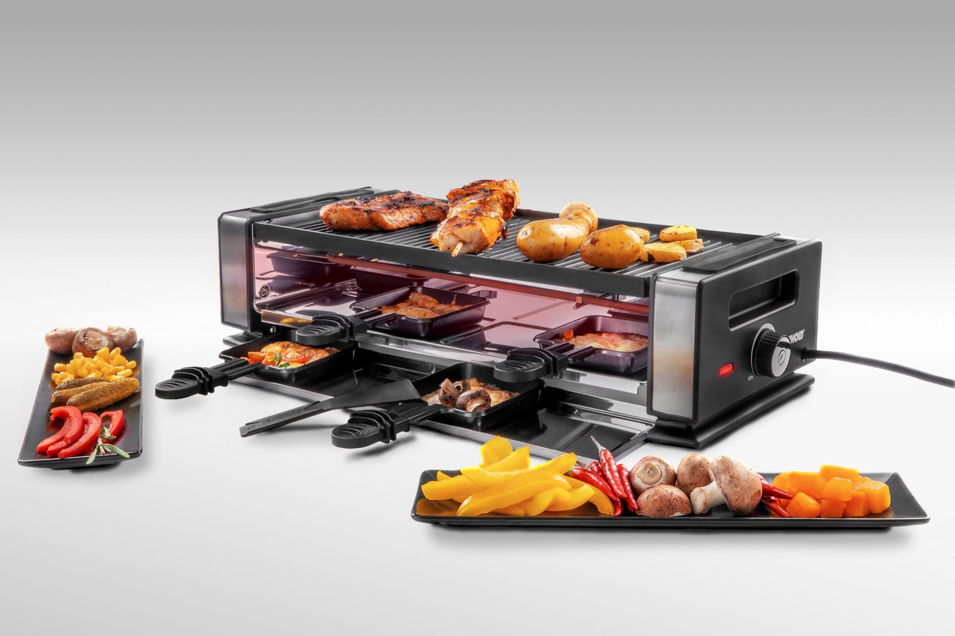 Das Unold Raclette Finesse Basic inklusive Raclette Buch besteht aus gekochtem Hähnchen, Kartoffeln und Gemüse auf der Oberseite, auf vier Tabletts befinden sich Käse und Gemüse. Davor stehen zwei schwarze Teller mit verschiedenen Gemüsescheiben.