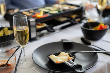 Eine Nahaufnahme eines Esstisches zeigt das Unold Raclette Finesse Basic, schwarze Teller, ein Glas Champagner, geschmolzenen Käse mit Gemüse und verschiedene Zutaten, die im Hintergrund arrangiert sind.