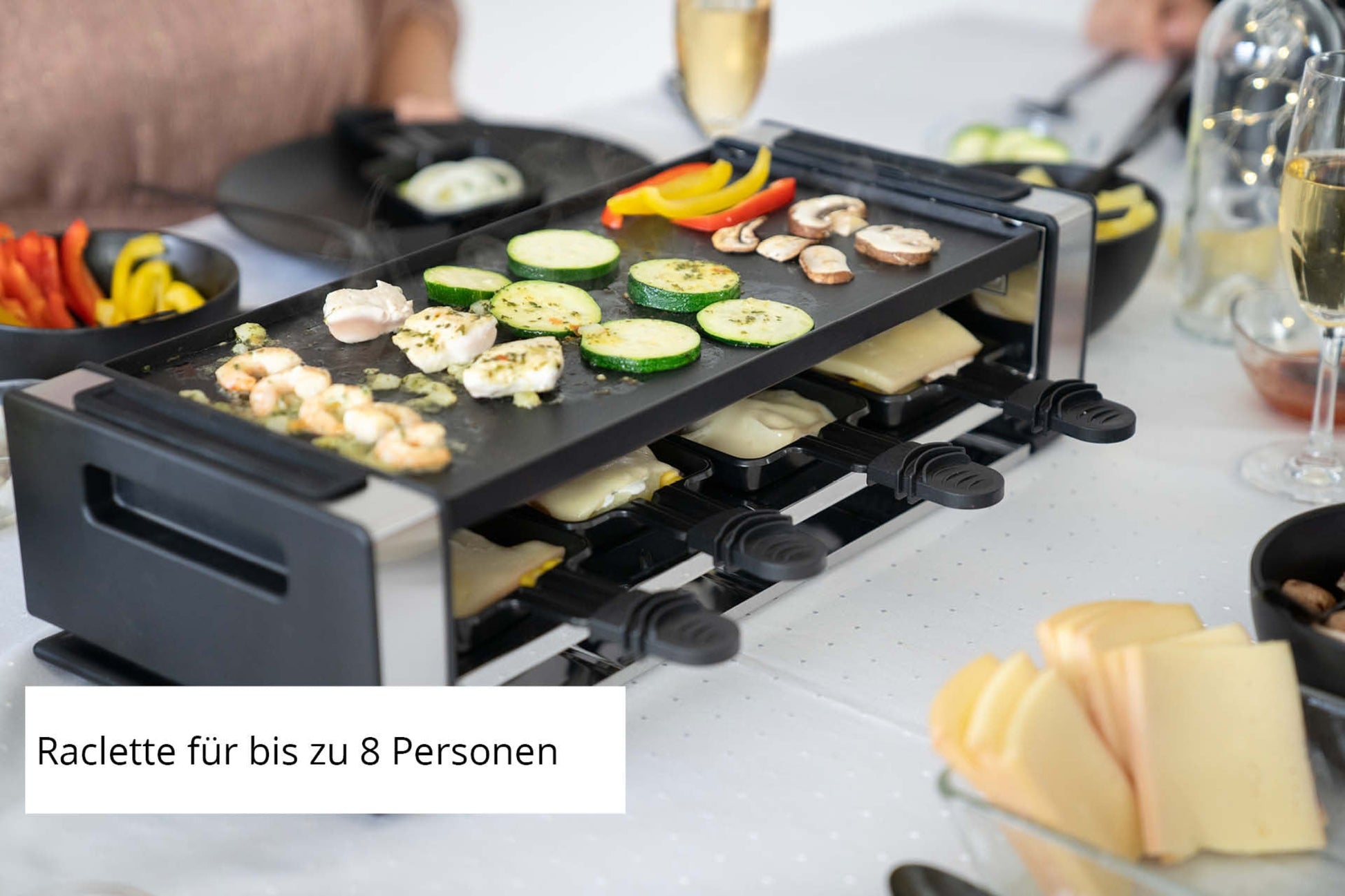Mit dem Unold Raclette Finesse Basic können bis zu 8 Personen Gemüse und Käse oben grillen, während kleine Tabletts darunter stehen. Teller mit Käsescheiben und Gemüse runden den Tisch ab. Bildunterschrift: Raclette für bis zu 8 Personen.