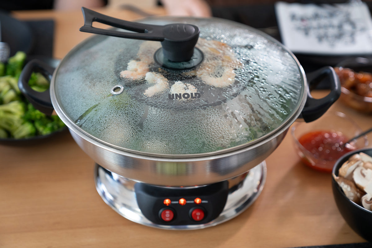 Im Unold Asia-Fondue, einem elektrischen Edelstahl-Hotpot mit Glasdeckel, werden Garnelen gedämpft. Teller mit Gemüse, Pilzen und Dip-Sauce stehen rund um den Topf auf einem Holztisch.
