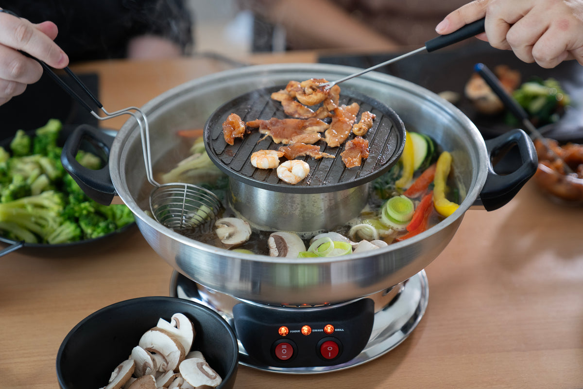 Zwei Personen kochen mit Utensilien Garnelen und Fleisch auf dem Unold Asia-Fondue Hot Pot Grill, umgeben von Gemüse, Pilzen und weiteren Zutaten, die rund um den Hot Pot auf dem Tisch angeordnet sind.