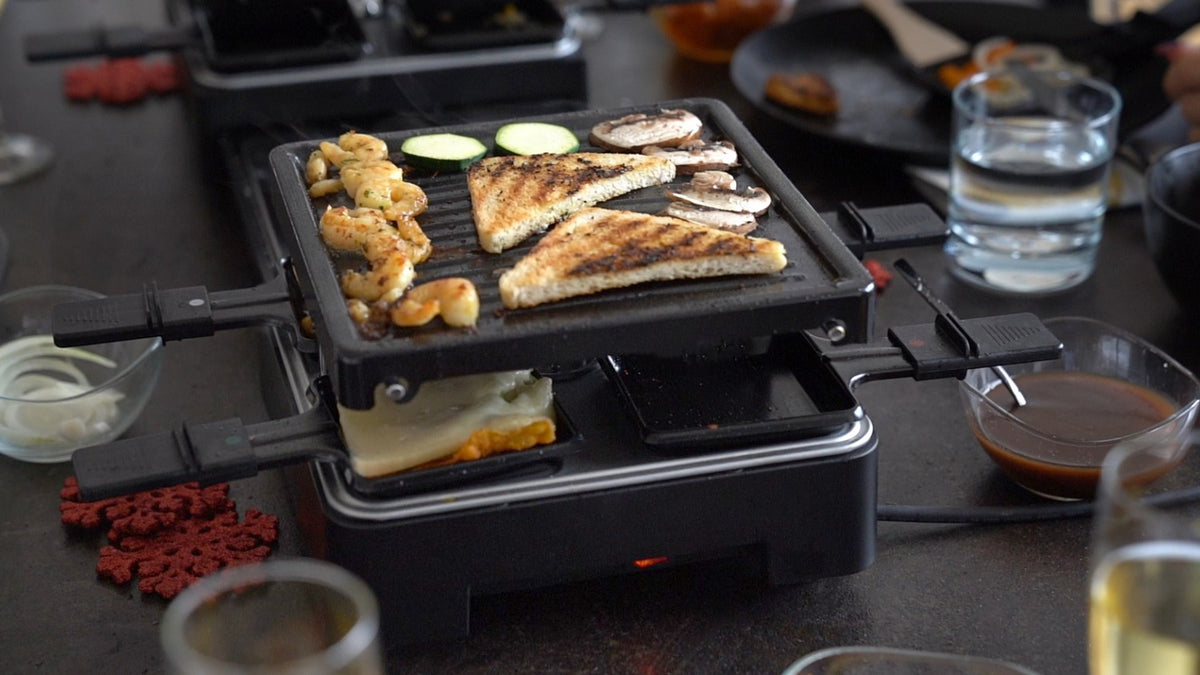 Auf dem ausziehbaren Raclette-Grill von Unold werden Brot, Garnelen, Zucchini und Pilze gegart, während in den darunterliegenden Schalen Käse schmilzt. Gläser mit Wasser und Geschirr stehen auf einem dunklen Tisch um den Grill herum.