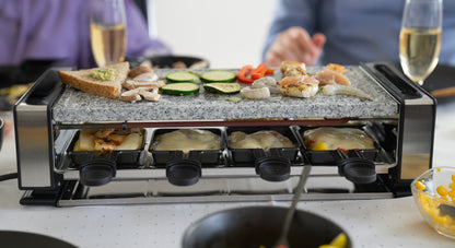 Das Unold Raclette Delice Basic grillt Gemüse, Garnelen und Brot obenauf, während der Käse unten schmilzt. Zwei Personen genießen das Essen am Tisch, im Hintergrund tönt ein Glas Weißwein.