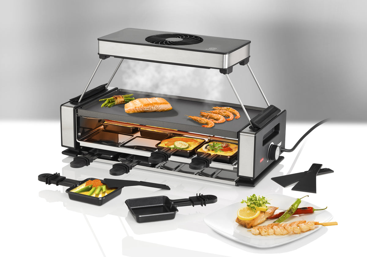 Das Unold Raclette Smokeless inklusive Raclette Buch verfügt über eine Metallplatte zum Grillen von Lachs, Garnelen und Gemüse. Unter den Tabletts schmelzen Käse und Beläge. Auf einem Beilagenteller befinden sich gegrillte Garnelen, Gemüse und eine Zitronengarnitur.
