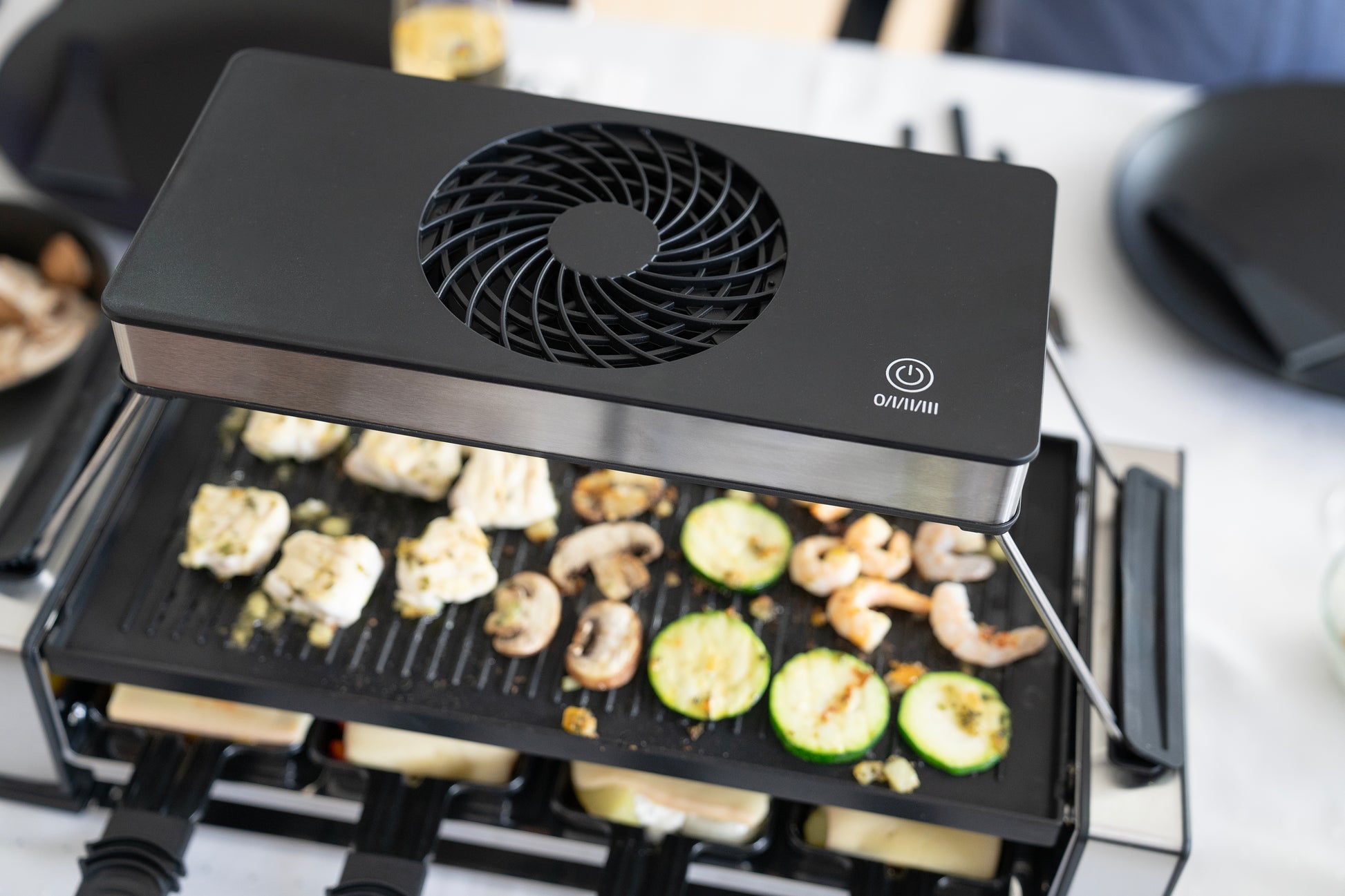 Der Unold Raclette Smokeless Tischgrill verfügt über einen Deckenventilator und gart Gemüse, Pilze, Garnelen und Fleischwürfel auf seiner Grillplatte; im Hintergrund sind schwarze Teller und Utensilien zu sehen.