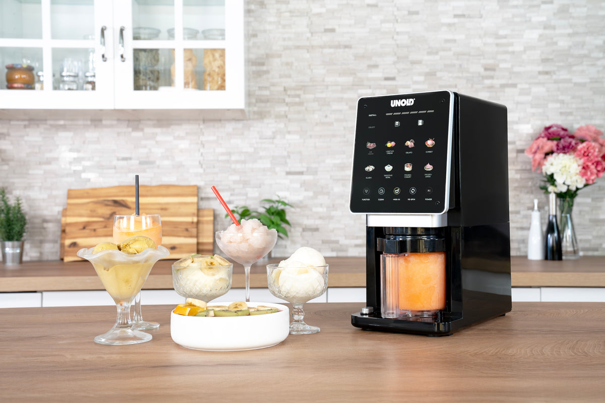 Die Unold AG Eismaschine Cream Dream steht auf einer modernen Küchentheke neben Desserts und Getränken - Eis, Smoothie und einem Getränk mit Strohhalm - mit Blumen und Schneidebrettern im Hintergrund.