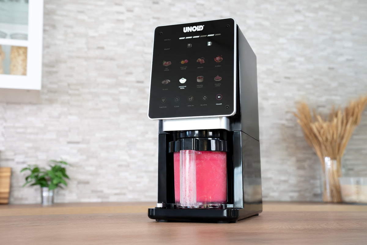 Die Unold AG Eismaschine Cream Dream, ein schwarzes Küchengerät mit Touchscreen und einem klaren Behälter mit leuchtend rosa Flüssigkeit, steht auf einer hölzernen Arbeitsplatte in einer modernen Küche.