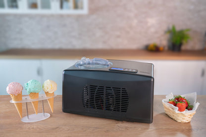 Die Unold Eismaschine Cortina 1,5 L steht auf einer Küchentheke neben einem Ständer mit drei Eistüten und einem Korb mit frischen Erdbeeren, im Hintergrund sind unscharfe Schränke und eine Pflanze zu sehen.