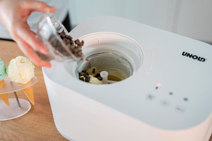 Eine Person füllt Schokoladenstückchen in eine mit Vanilleeis gefüllte Unold Eismaschine Snow 1 L. Daneben stehen drei Waffeln mit grünen und gelben Kugeln auf einem Teller. Die weiße Maschine trägt die Marke Unold.