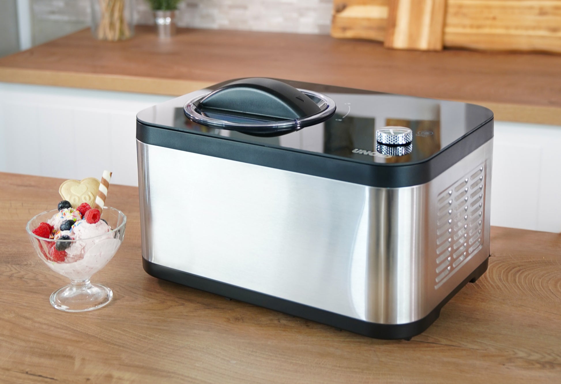 Die Unold Eismaschine & Joghurtbereiter EVA steht auf einer hölzernen Arbeitsplatte neben einer Glasschale mit Eis, Beeren und einer Waffel in einer hellen, modernen Küche.