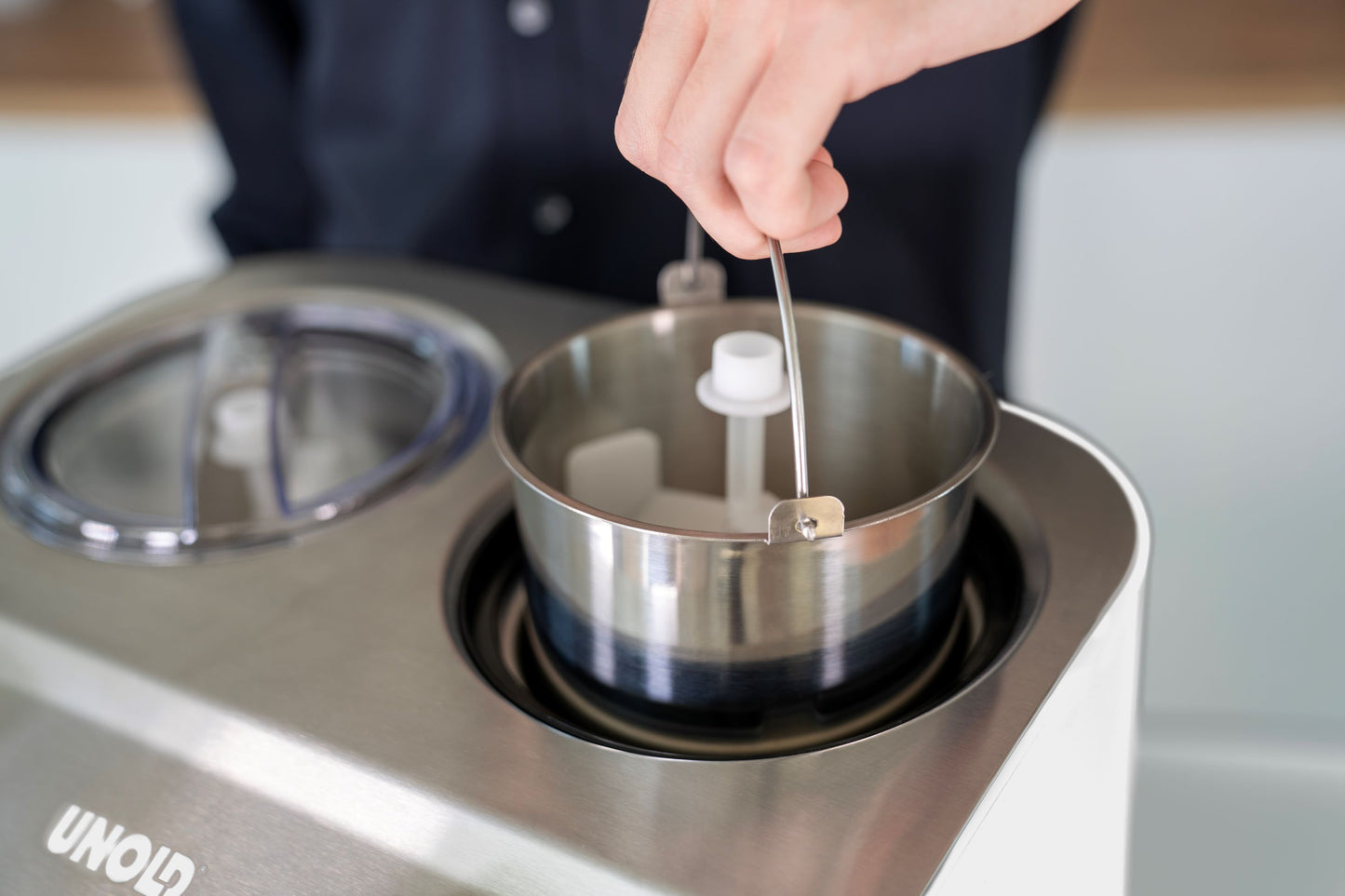 Eine Person stellt eine Metallrührschüssel mit Griff in die Unold EISMASCHINE Doppio 2,4 L-Eismaschine, die auf einer Küchentheke steht, oder nimmt sie heraus.