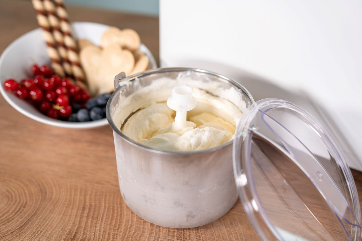 Die Unold EISMASCHINE Doppio 2,4 L, gefüllt mit Vanilleeis, steht offen auf einer Holzplatte. Daneben eine Schale mit Johannisbeeren, Heidelbeeren, Keksen und Waffelröllchen.
