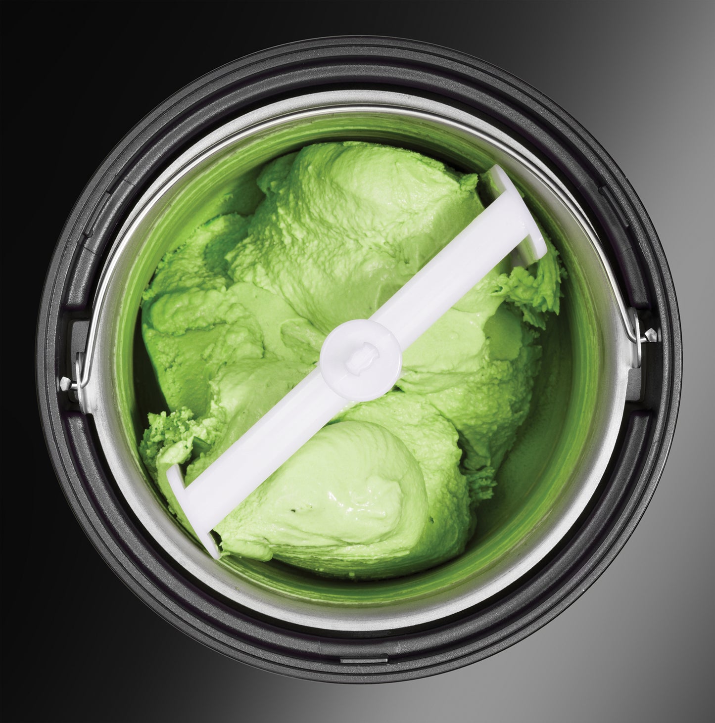 Draufsicht auf eine grüne Eismischung in der Unold Eismaschine Gusto inklusive Eisbuch 2 L, mit einem weißen Paddel in der Mitte, vor einem dunklen Hintergrund.