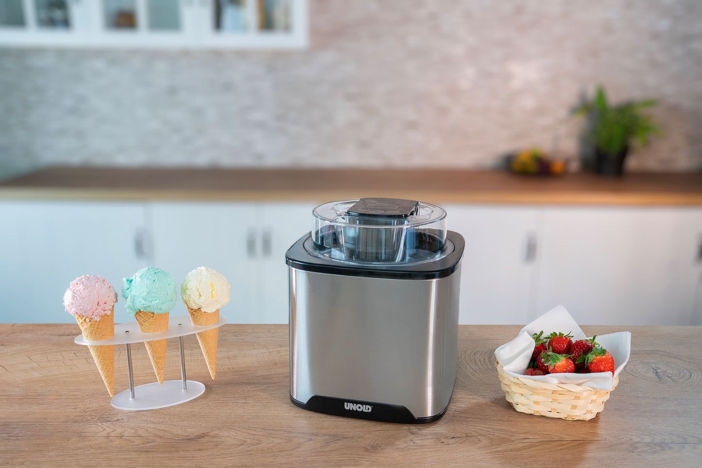 Die Eismaschine „Unold Eismaschine Gelato 1,5 L“ steht auf einer Küchentheke neben einem Halter mit drei Eistüten und einem Korb mit frischen Erdbeeren, im Hintergrund sind weiße Schränke und eine Rückwand zu sehen.
