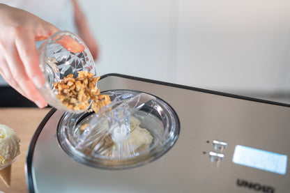 Eine Person schüttet gehackte Walnüsse aus einer kleinen Glasschale in eine Unold Eismaschine One 1,2 L, die eine teilweise gemischte Eiscremebasis enthält, während die Digitalanzeige der Maschine sichtbar ist.