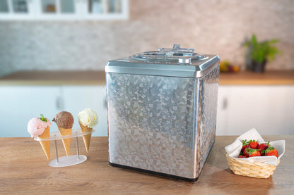 Die Unold Eismaschine Profi 2 L steht auf einer Küchentheke neben einem Ständer mit drei Eistüten und einem Korb mit frischen Erdbeeren, im Hintergrund ist eine helle, verschwommene Küche zu sehen.