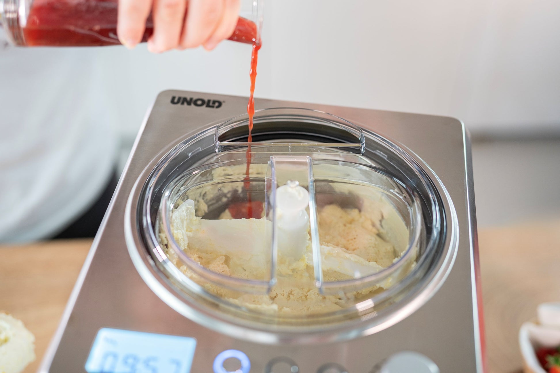 Nahaufnahme: Eine Person gießt rote Flüssigkeit in die Unold Eismaschine Profi 2L, die eine helle Mischung enthält. Die Maschine verfügt über eine Digitalanzeige und der Deckel ist teilweise geöffnet.