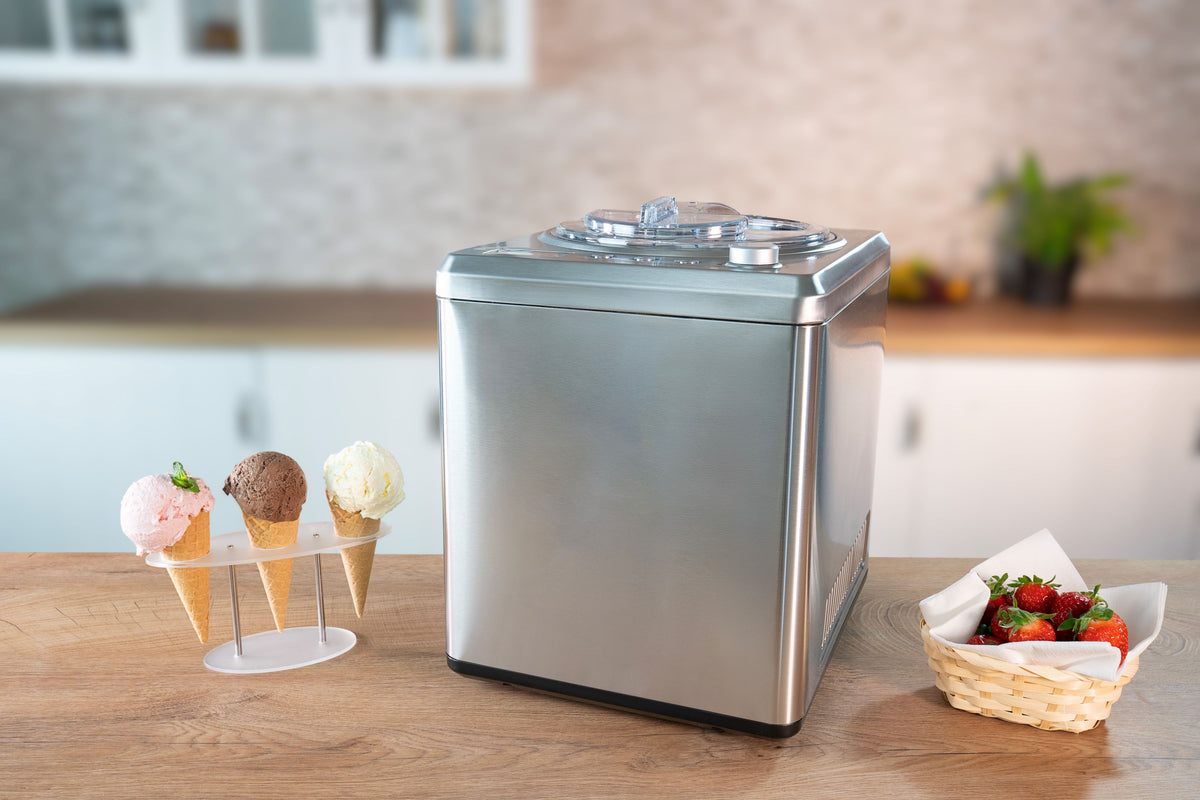 Die Unold Eismaschine Exklusiv 2 L aus Edelstahl steht auf einer Küchentheke neben drei Eistüten in einem Ständer und einem Korb mit Erdbeeren vor einem unscharfen Küchenhintergrund.