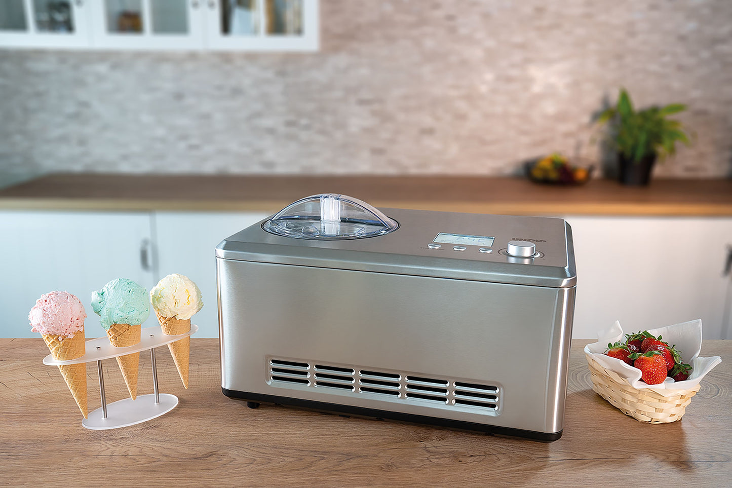Die Unold B-Ware Eismaschine Nobile 2 L steht auf einer Küchentheke neben einem Ständer mit vier Eistüten und einem kleinen Korb Erdbeeren vor einem modernen Küchenhintergrund.