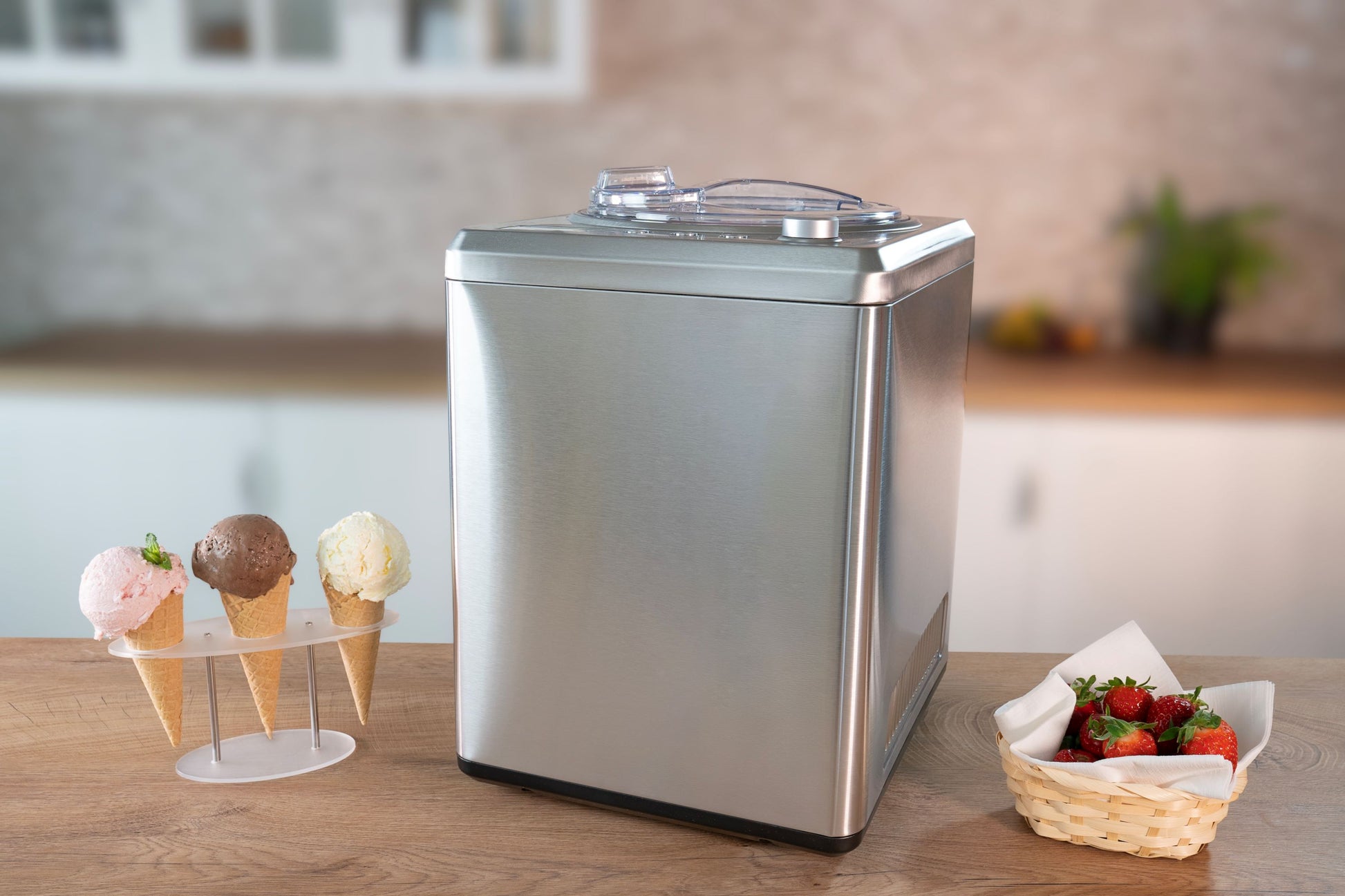 Die Unold Eismaschine Profi Plus 2,5 L steht auf einer Küchentheke neben drei Eistüten auf einem Ständer und einem Korb mit frischen Erdbeeren, im Hintergrund ist eine verschwommene moderne Küche zu sehen.