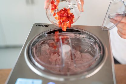 Eine Person schüttet gehackte Erdbeeren aus einer Schüssel in die Unold Eismaschine Profi Plus 2,5 L, die eine rosafarbene gefrorene Mischung enthält.
