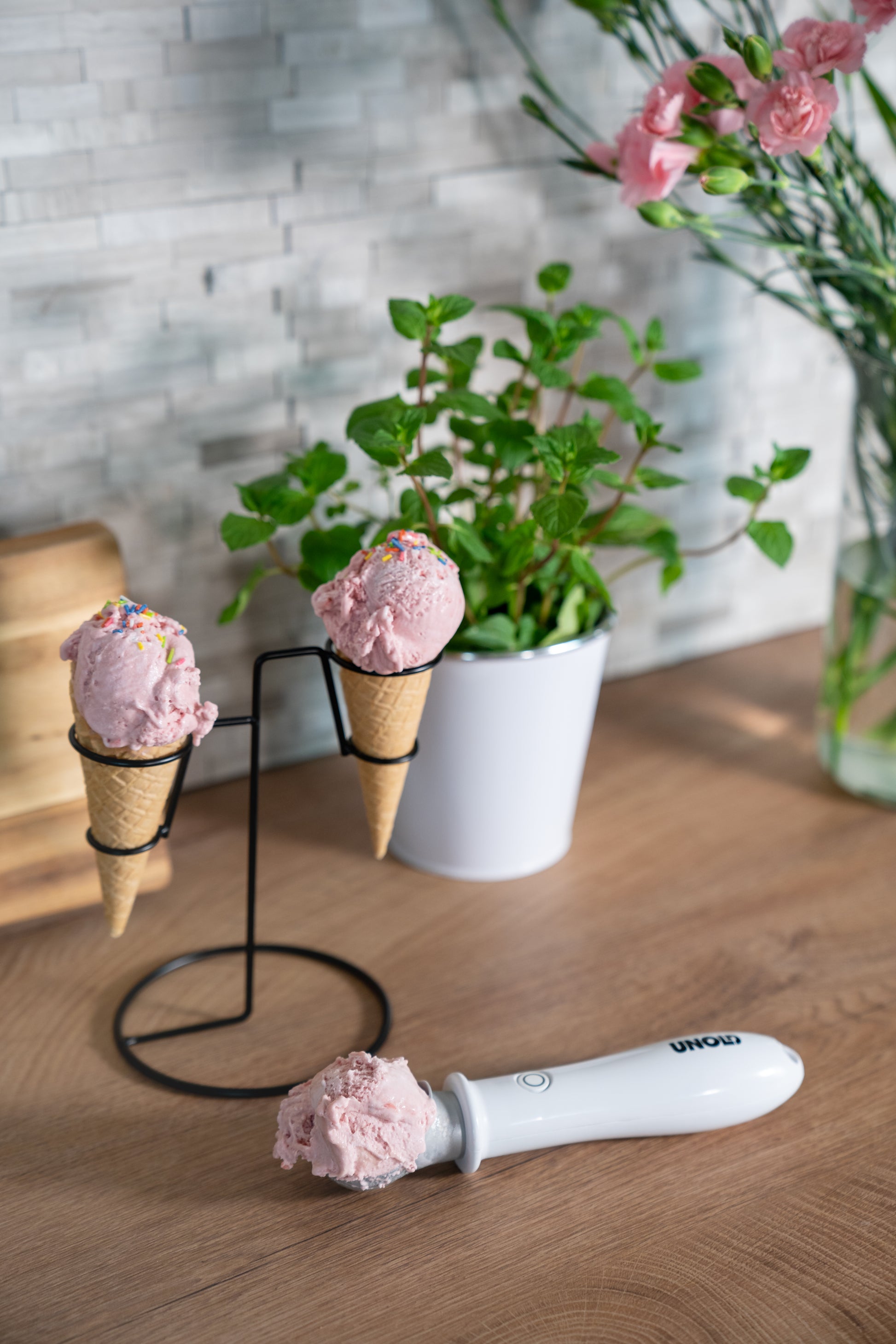 Drei Kugeln rosa Eis, zwei in Waffeln auf einem Metallständer und eine auf dem Beheizten Eisportionierer Carlo von Unold, liegen auf einer Holzoberfläche mit einer Topfpflanze und rosa Blumen im Hintergrund.