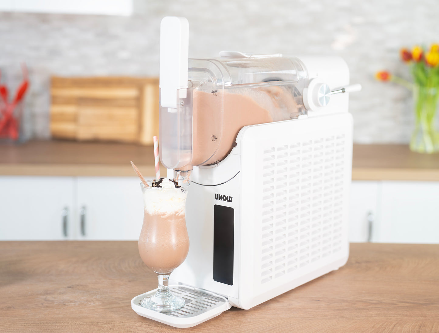 Die weiße Unold Slush-Eismaschine Susi gibt einen Schokoladenmilchshake mit Schlagsahne in ein hohes Glas mit gestreiftem Strohhalm und Löffel aus, das auf einer Küchentheke vor einem modernen Küchenhintergrund steht.
