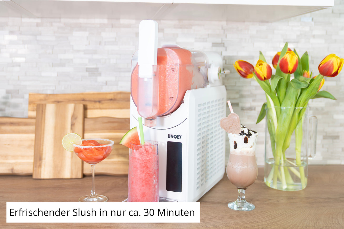 Die Unold Slush-Eismaschine Susi steht auf der Küchentheke und spritzt roten Slush in ein Glas. Daneben stehen zwei Getränke – ein rotes mit Limette, ein braunes mit Schlagsahne – sowie eine Vase mit Tulpen und Schneidebrettern im Hintergrund.