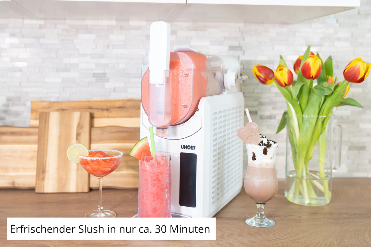 Die Unold Slush-Eismaschine Susi steht auf der Küchentheke und spritzt roten Slush in ein Glas. Daneben stehen zwei Getränke – ein rotes mit Limette, ein braunes mit Schlagsahne – sowie eine Vase mit Tulpen und Schneidebrettern im Hintergrund.