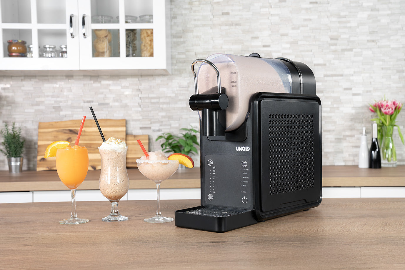 Die Unold Slush-Eismaschine Stella, eine schwarze Thekengetränkemaschine, schenkt cremige Getränke in drei Gläser mit Strohhalmen und Orangenscheiben auf einer hölzernen Küchentheke mit weißen Schränken und einer Blumenvase im Hintergrund aus.