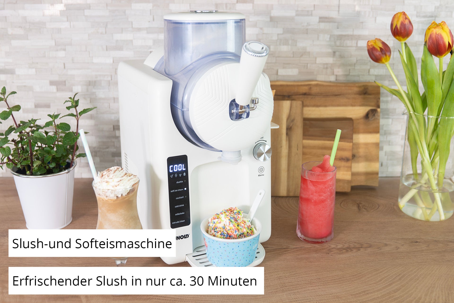 Die Unold 2in1-Slush- und Softeismaschine Sofie steht auf einer Küchentheke. Im Vordergrund stehen zwei bunte Getränke – ein rotes Slush-Getränk und ein Softeis mit Streuseln. Im Hintergrund sind Tulpen, Utensilien und ein deutscher Text zu sehen, der die Maschine beschreibt.